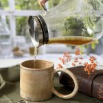Main versant du thé avec une carafe dans le mug GARANCE en grès artisanal, posé sur une table de cuisine avec un livre et une cuillère à côté.