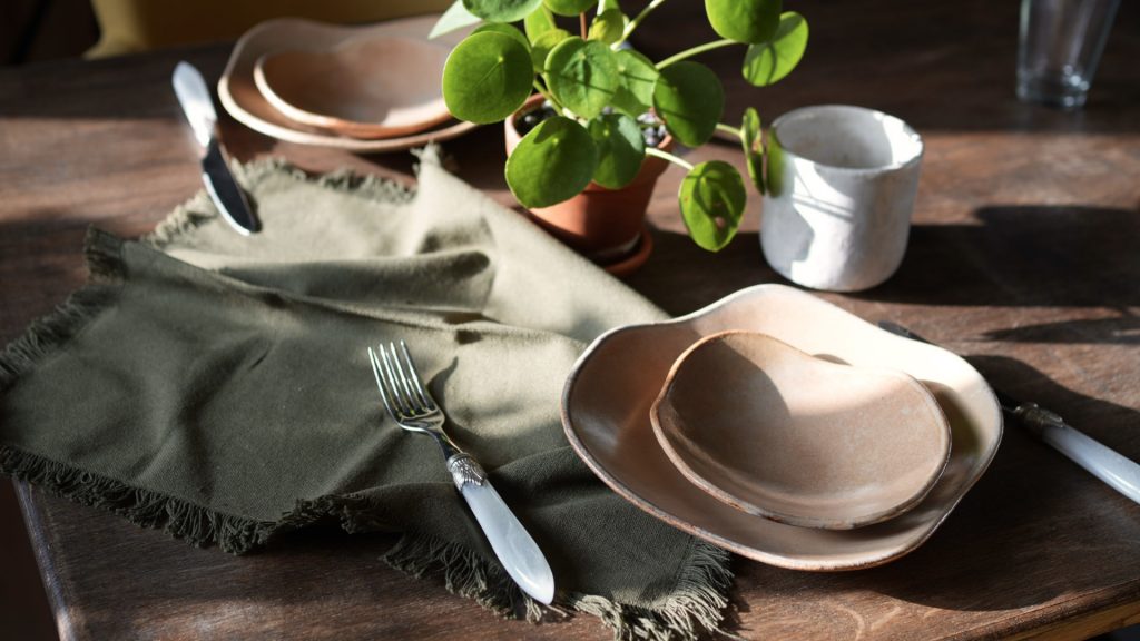 Mise en scène chaleureuse d'une table dressée avec les assiettes LINA en grès artisanal et les coupelles MILA de LIS Céramique, accompagnées de couverts, d'une petite plante et d'une serviette, prête pour un repas.