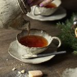 Théière Cosmos en grès artisanal qui verse du thé dans la tasse lilium sur la sous-tasse Dahlia avec la cuillère rosa posées sur une table dressée.