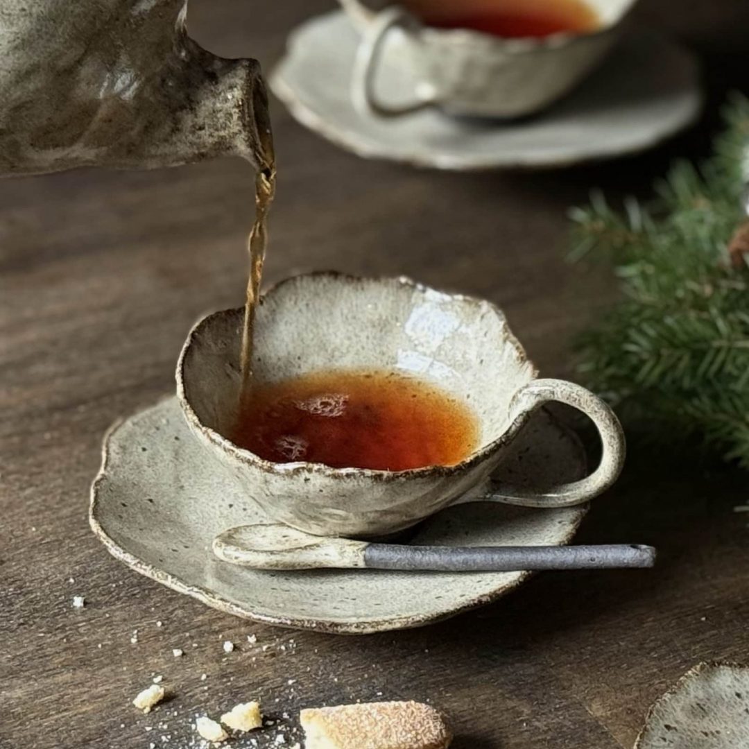 Tasse Lilium en grès artisanal posée sur la sous-tasse Dahlia avec la cuillère Rosa, service de thé avec la théière Cosmos sur une table, misse en situation avec biscuits et autres tasses en arrière-plan.