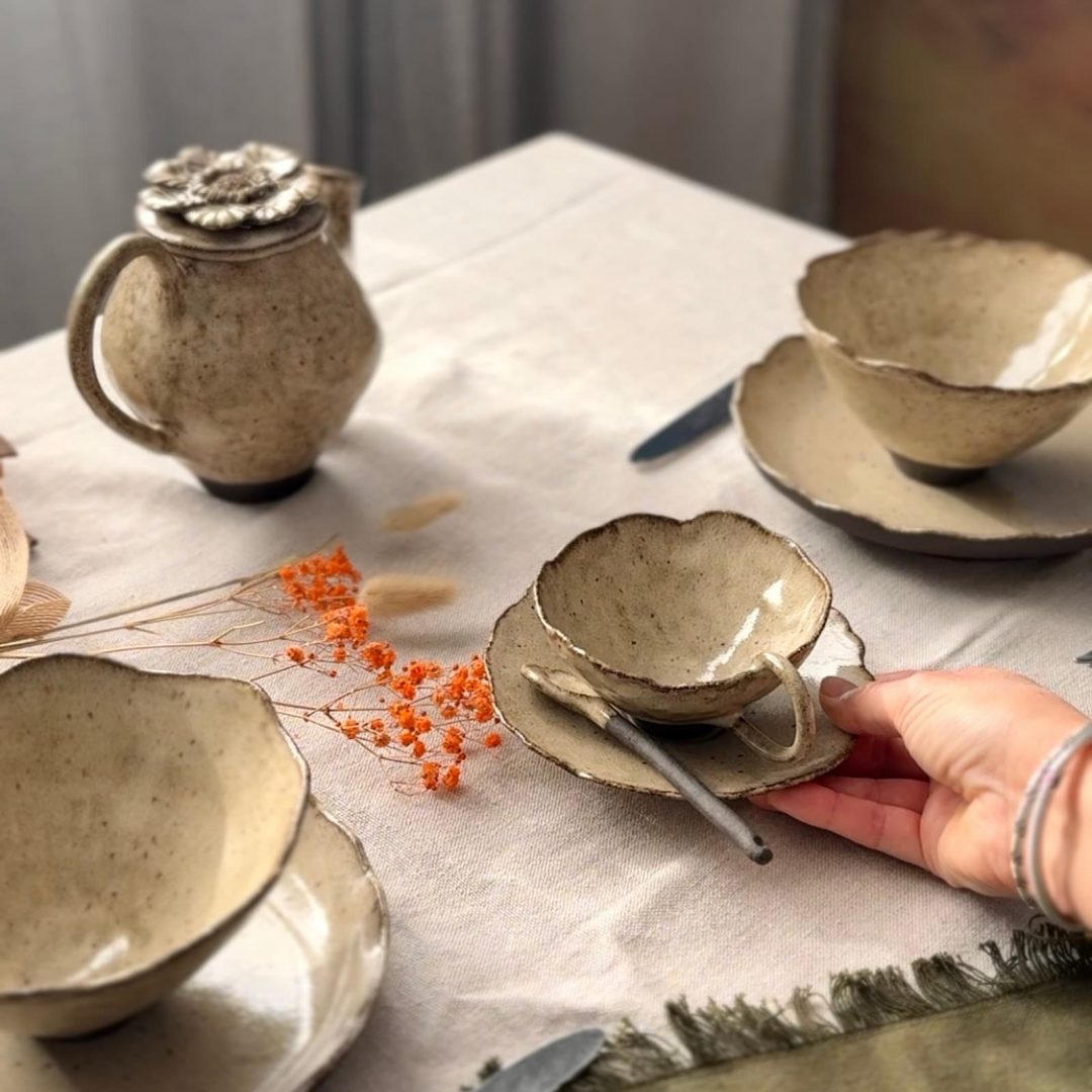 Main posant une tasse avec sous-tasse et cuillère sur une table dressée, avec théière Cosmos, bol Camellia en grès artisanal et assiette Jasmina en arrière plan.