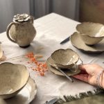 Main posant une tasse avec sous-tasse et cuillère sur une table dressée, avec théière Cosmos, bol Camellia en grès artisanal et assiette Jasmina en arrière plan.