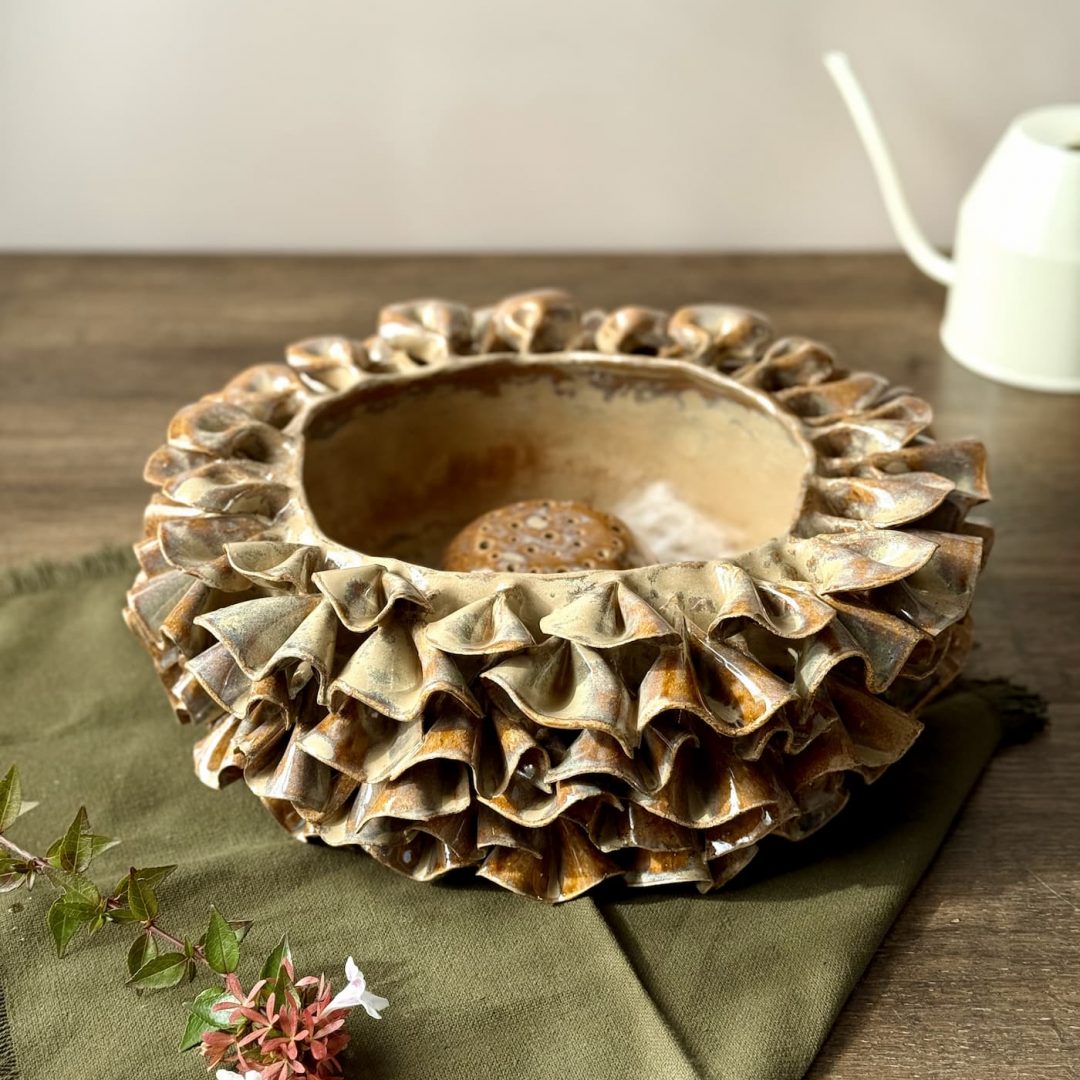 Vase ÉMILE en céramique artisanale posé sur une table avec son dôme perforé visible, une fleur à côté et un arrosoir en arrière-plan.