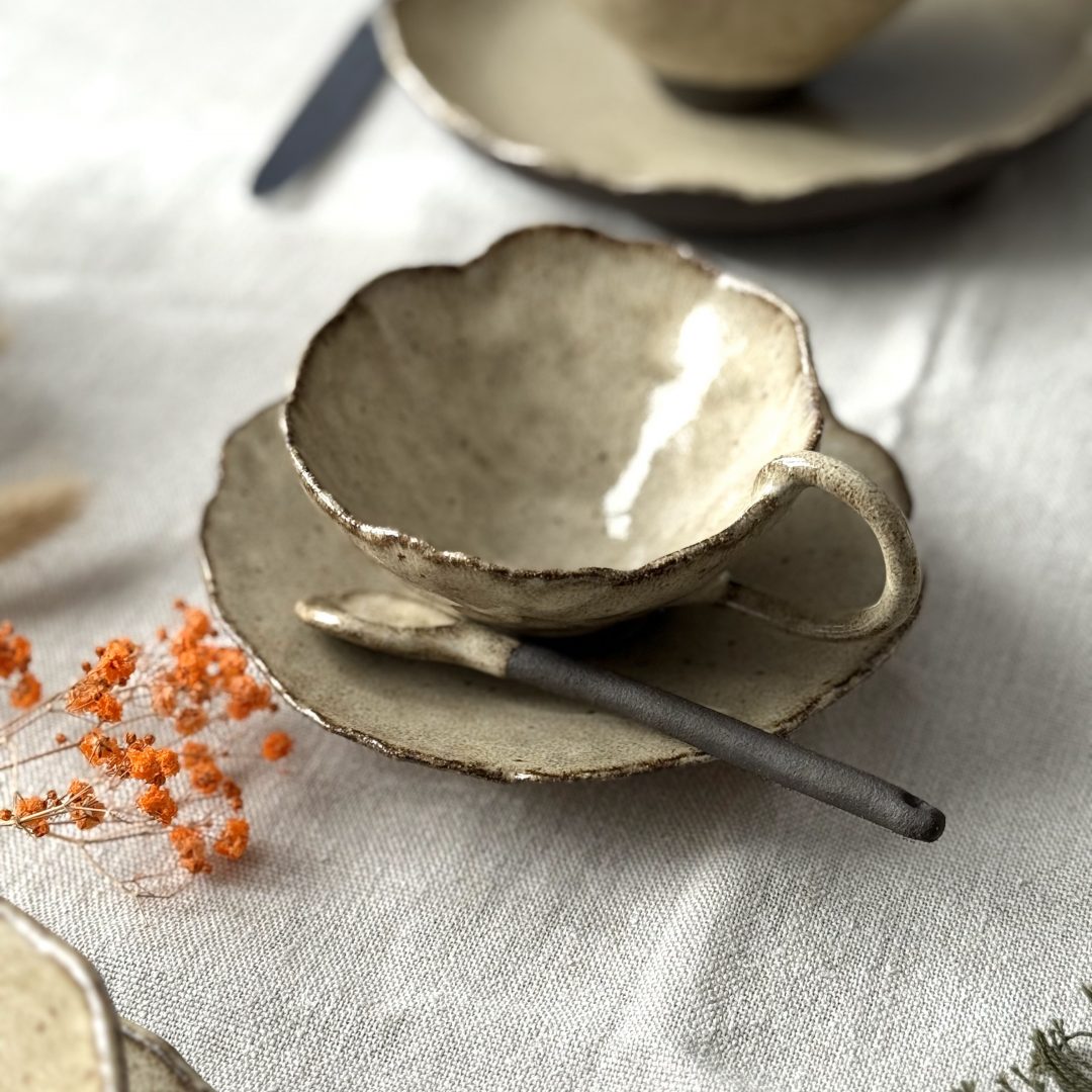 Tasse Lilium en grès noir de profil posée sur la sous-tasse Dahlia avec la cuillère Rosa, en mise en scène sur une table, avec l’assiette Jasmina en arrière-plan.
