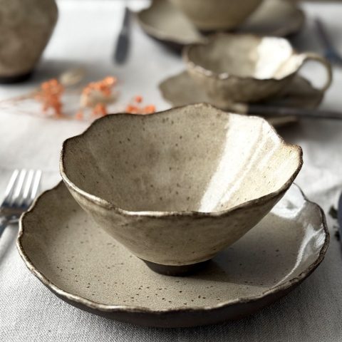 Bol Camellia en céramique artisanale posé sur l’assiette Jasmina au premier plan, avec la tasse Lilium, la sous-tasse Dahlia et la cuillère Rosa en arrière-plan sur la table dressée.