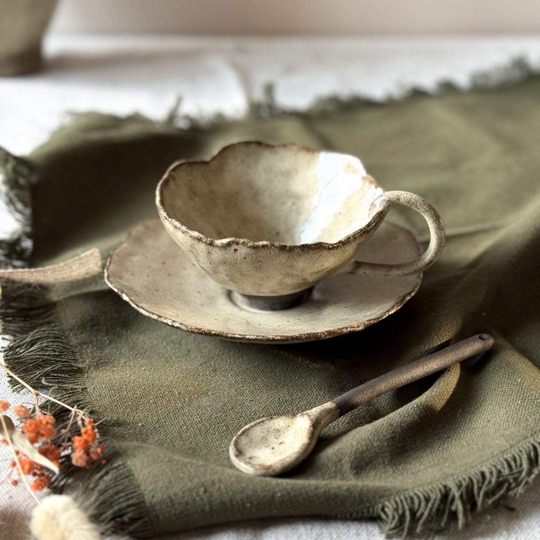 Tasse Lilium en céramique artisanale vue en légère plongée, posée sur la sous-tasse Dahlia avec la cuillère Rosa, sur une table.