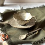 Tasse Lilium en céramique artisanale vue en légère plongée, posée sur la sous-tasse Dahlia avec la cuillère Rosa, sur une table.