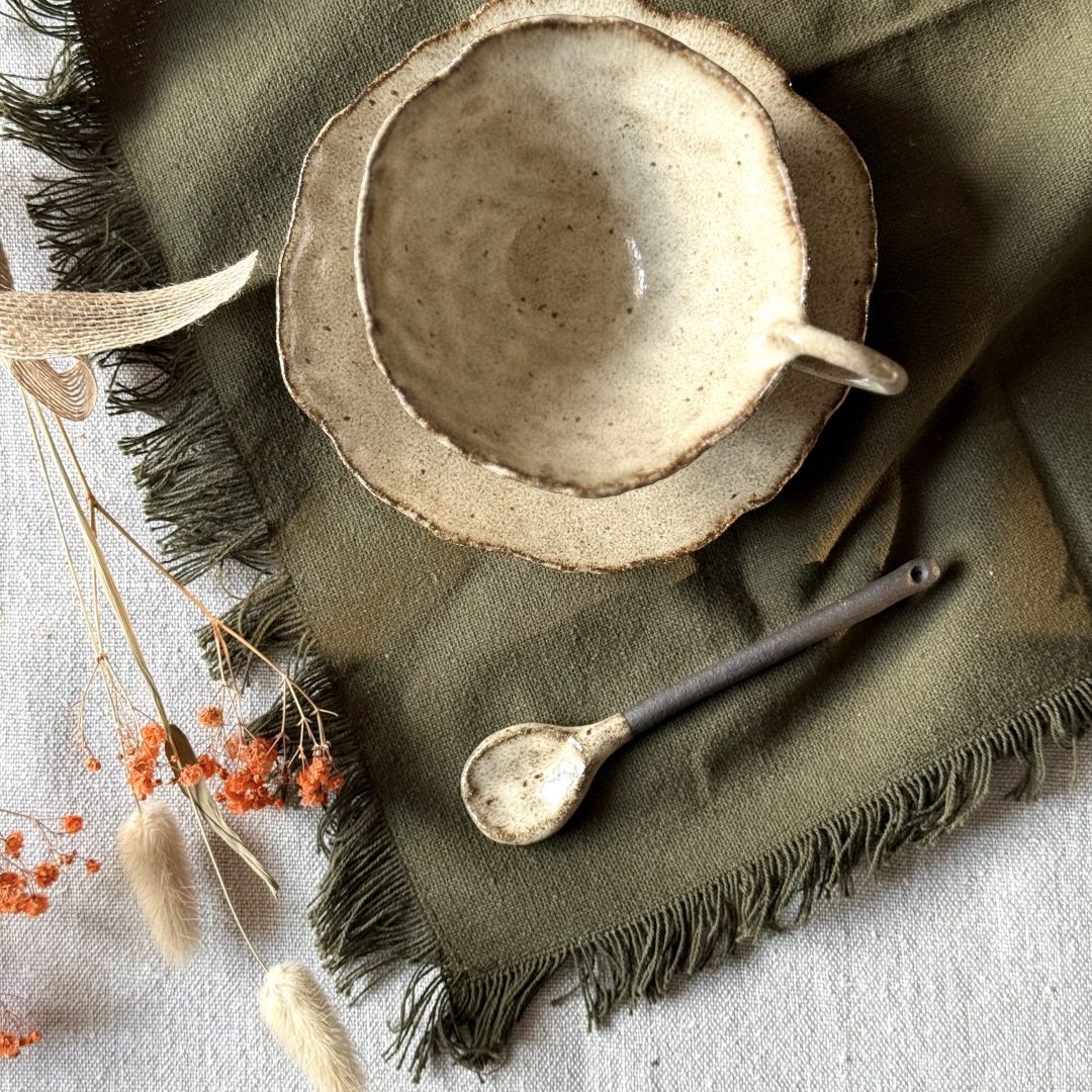 Tasse Lilium en grès noir émaillé crème vue de dessus, posée sur la sous-tasse Dahlia avec la cuillère Rosa, sur une table en lin.