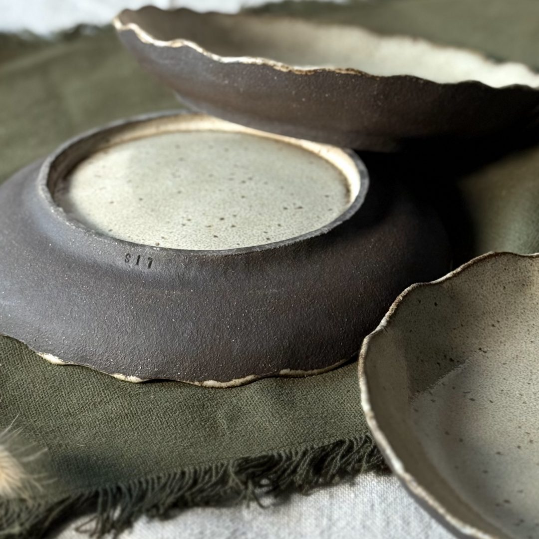 Trois assiettes Jasmina posées sur la table, dont une retournée montrant le pied émaillé, la signature sur le bord et la terre noire visible sur les bords des assiettes.