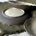 Trois assiettes Jasmina posées sur la table, dont une retournée montrant le pied émaillé, la signature sur le bord et la terre noire visible sur les bords des assiettes.