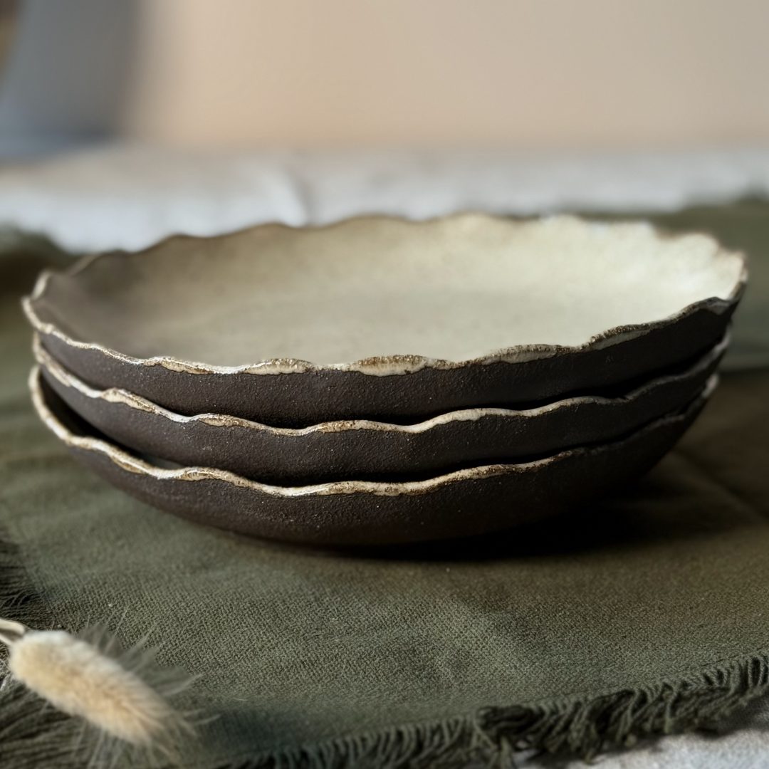 Trois assiettes Jasmina empilées, vues de profil, montrant la terre noire et le bord délicatement émaillé, posées sur une table.