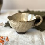 Tasse Aster en grès noir émaillé crème, vue de profil et posée sur une table.