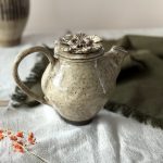 Théière cosmos en grès noir émaillé crème, vue de profil et posée sur une table.