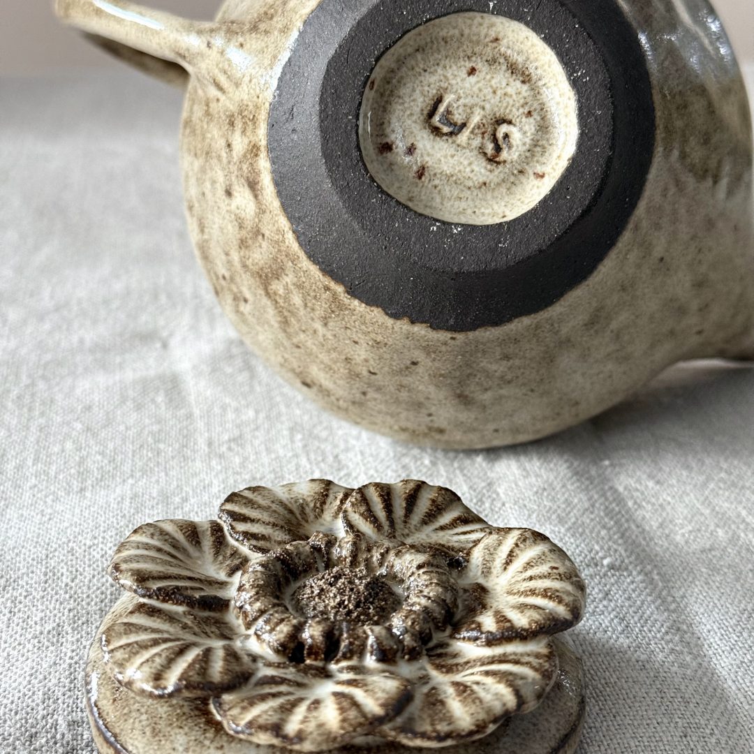 Théière Cosmos couchée montrant le dessous émaillé et la signature, avec le couvercle sculpté en forme de fleur posé devant, le tout en céramique artisanale en grès noir.