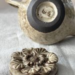 Théière Cosmos couchée montrant le dessous émaillé et la signature, avec le couvercle sculpté en forme de fleur posé devant, le tout en céramique artisanale en grès noir.