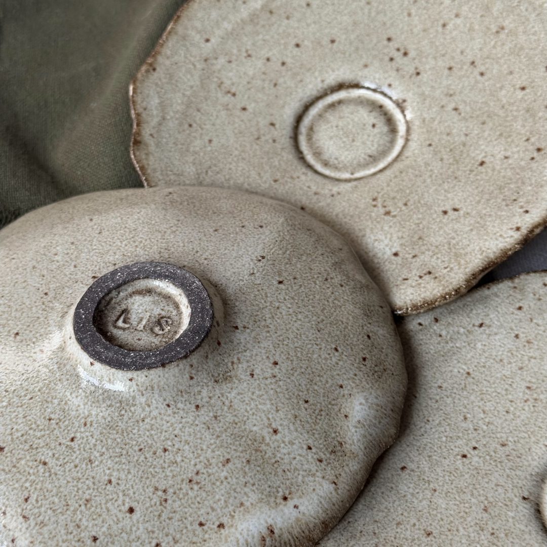 Sous-tasse Dahlia en grès noir retournée montrant le dessous signé et le renfort central, avec deux autres sous-tasses posées à côté.