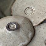 Sous-tasse Dahlia en grès noir retournée montrant le dessous signé et le renfort central, avec deux autres sous-tasses posées à côté.