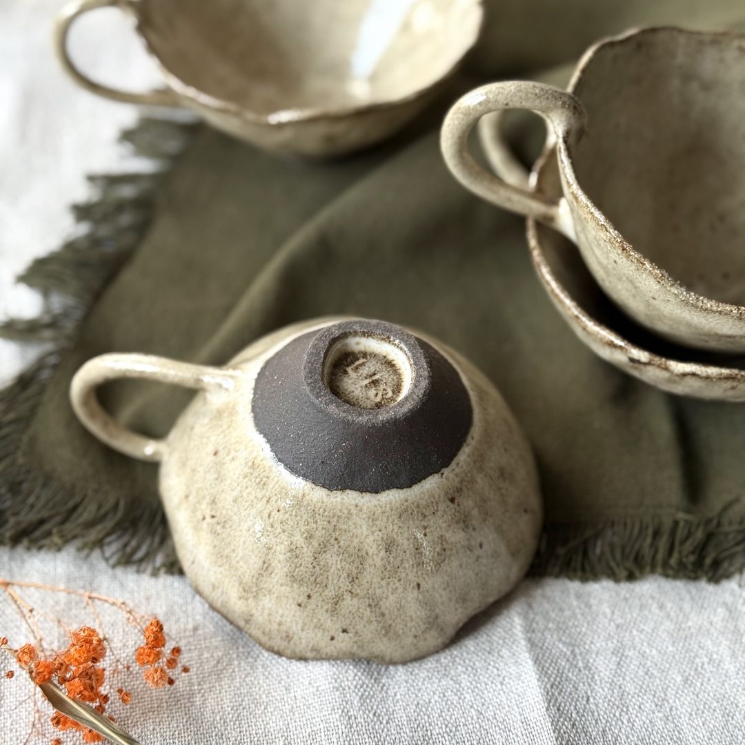 Dessous de la tasse Lilium retournée avec la signature de Lis Céramique, posée sur une table, avec d’autres tasses Lilium en grès noir empilées en arrière-plan.