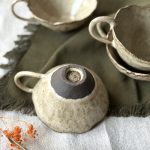 Dessous de la tasse Lilium retournée avec la signature de Lis Céramique, posée sur une table, avec d’autres tasses Lilium en grès noir empilées en arrière-plan.