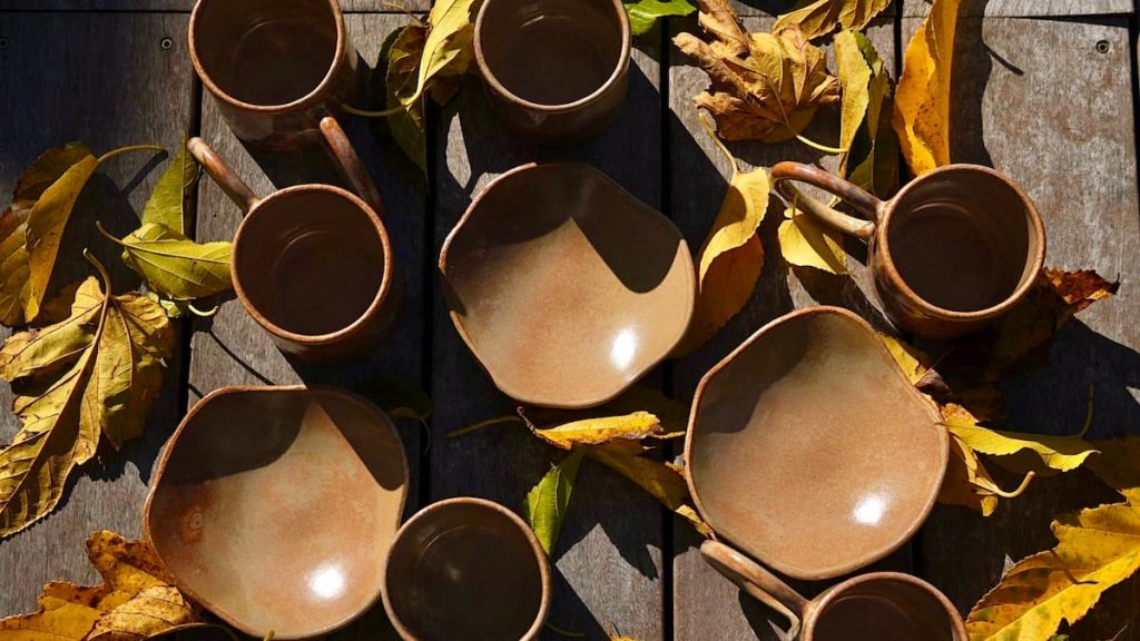Nuances beiges et dorées : coupelles MILA et mugs GARANCE tout juste sortis du four, posés sur la terrasse parmi les feuilles d'automne.