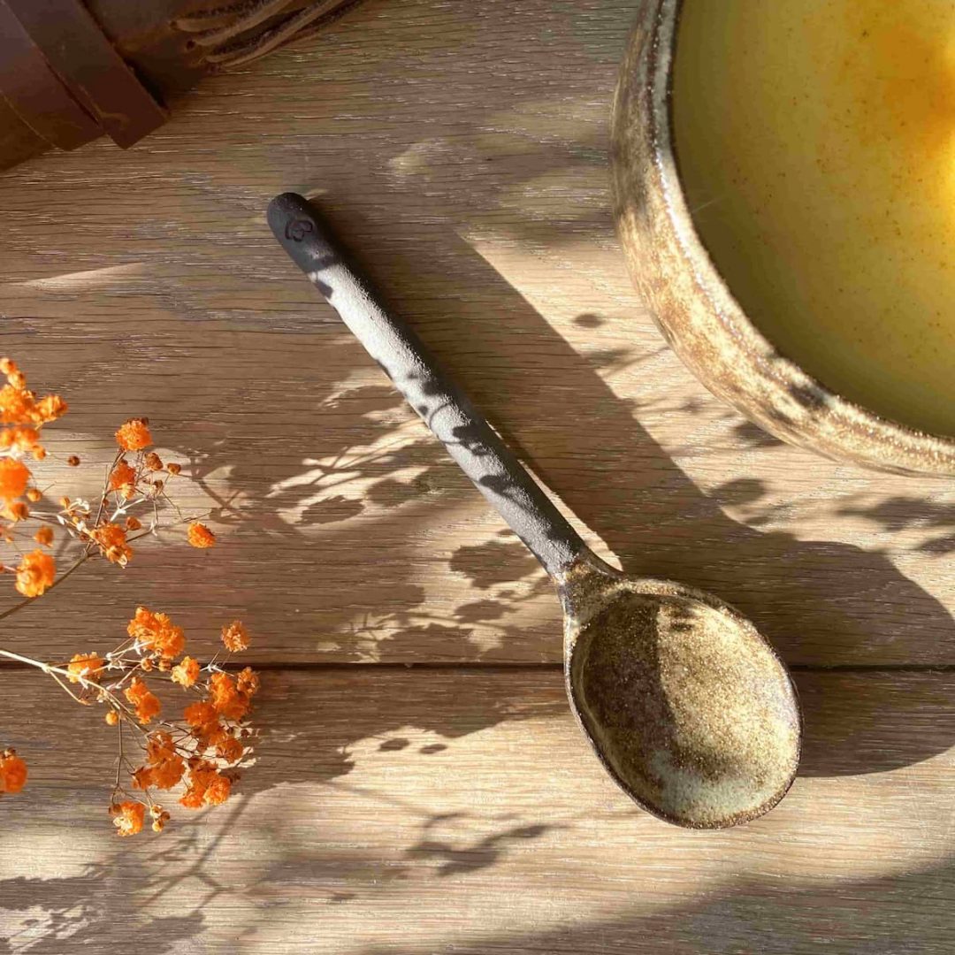 Cuillère Bintou en grès façonnée à la main, posée près d'un bol de soupe, éclairée par une lumière naturelle.