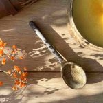 Cuillère Bintou en grès façonnée à la main, posée près d'un bol de soupe, éclairée par une lumière naturelle.