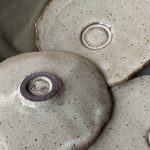 Sous-tasse Dahlia en grès noir retournée montrant le dessous signé et le renfort central, avec deux autres sous-tasses posées à côté.