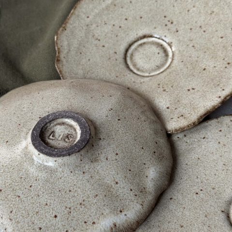 Sous-tasse Dahlia en grès noir retournée montrant le dessous signé et le renfort central, avec deux autres sous-tasses posées à côté.