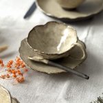 Tasse Lilium en grès noir de profil posée sur la sous-tasse Dahlia avec la cuillère Rosa, en mise en scène sur une table, avec l’assiette Jasmina en arrière-plan.