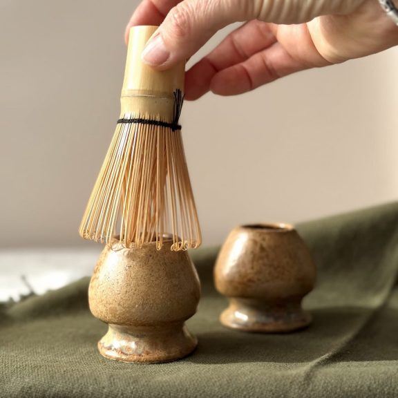 Main posant un fouet matcha sur le support à fouet matcha Nori en grès blanc, avec un second fouet matcha en arrière-plan.