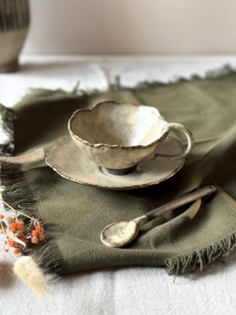 Tasse Lilium en céramique artisanale vue en légère plongée, posée sur la sous-tasse Dahlia avec la cuillère Rosa, sur une table.